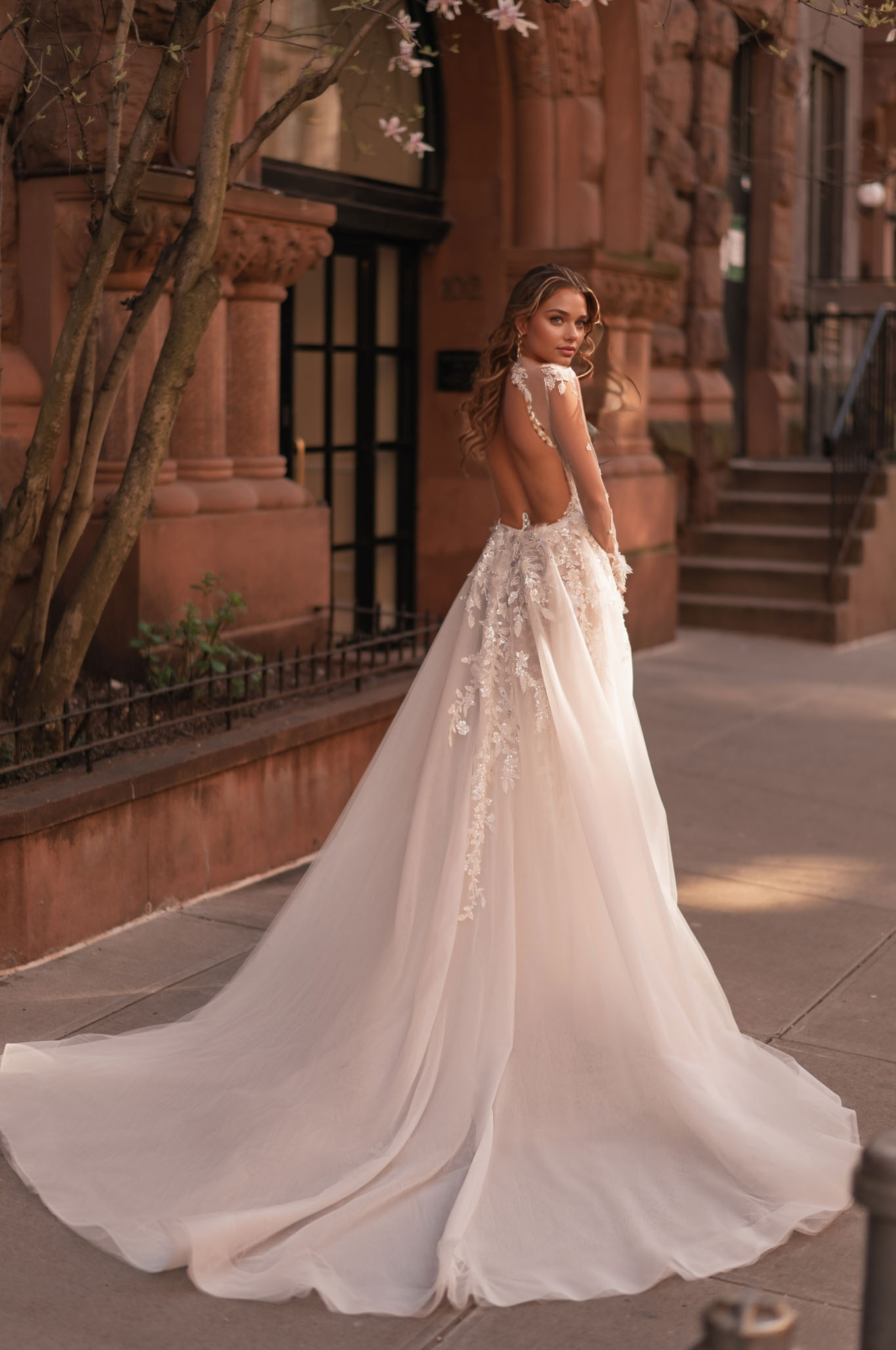 woman wearing a floral appliqué long-sleeve sheer illusion wedding dress with long train by a classic building
