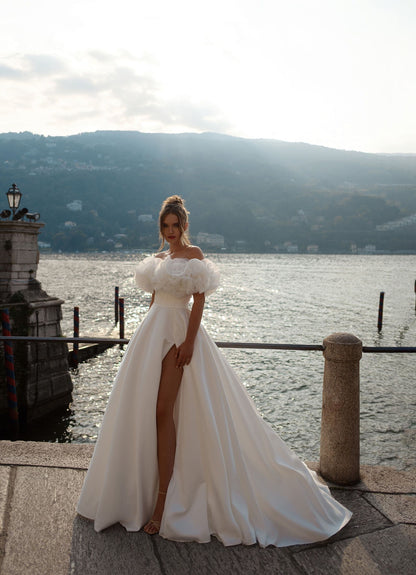 Modern princess-style wedding gown featuring an off-shoulder neckline, leg-revealing high slit, and couture-level floral sleeve accents.