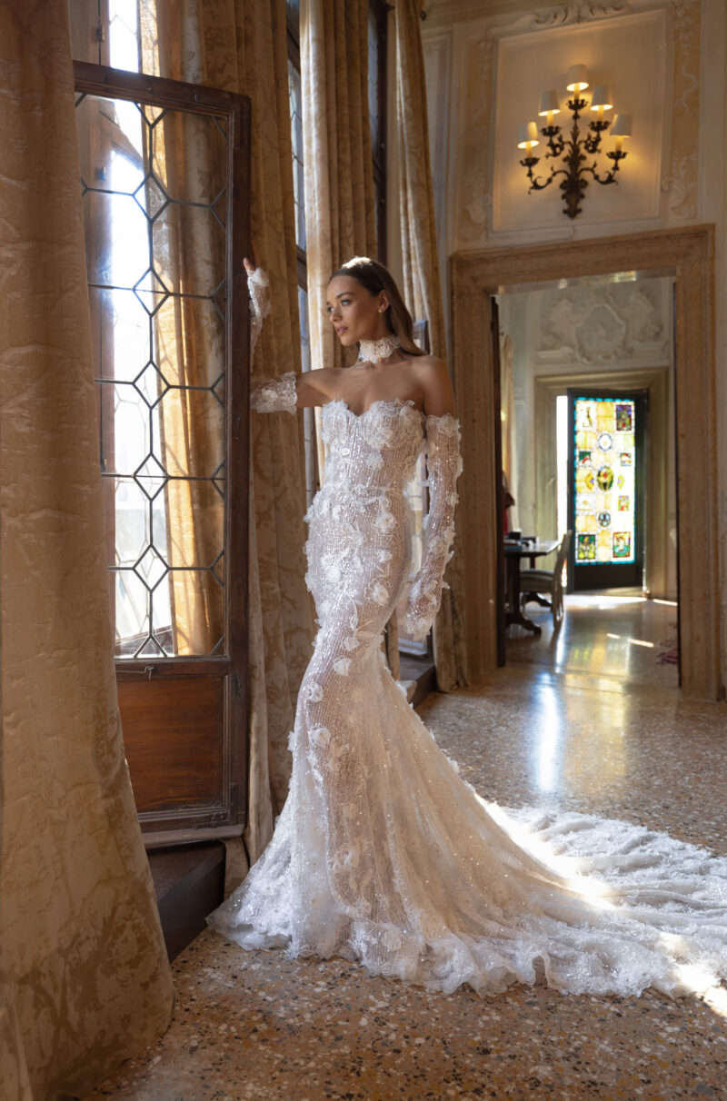 Bride in glowing 3D lace mermaid gown standing by window